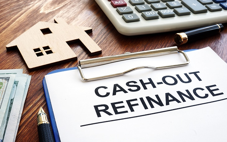 Clipboard with sheet of paper that says "cash-out refinance" sits on a desk with a calculator and wood house cutout.